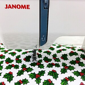 May include: A white sewing machine with the word "JANOME" printed in red on the side. The machine is sewing on a white fabric with a green and red holly pattern.
