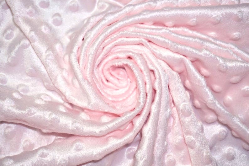 Op de afbeelding: Een close-up van een zachte, roze stof met een textuur, gestippeld patroon. De stof is gevouwen en gekreukt, waardoor een wervelend effect ontstaat.
