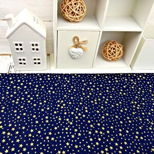 May include: A navy blue fabric with a repeating pattern of small, yellow stars. The fabric is displayed on a white wooden surface, with a white decorative house and a white shelf unit in the background.