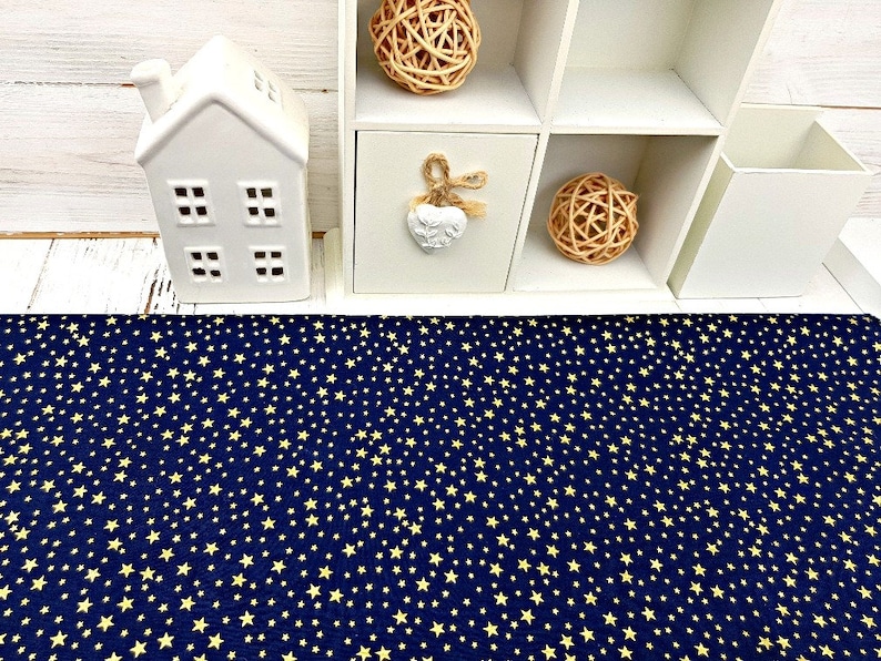 May include: A navy blue fabric with a repeating pattern of small, yellow stars. The fabric is displayed on a white wooden surface, with a white decorative house and a white shelf unit in the background.