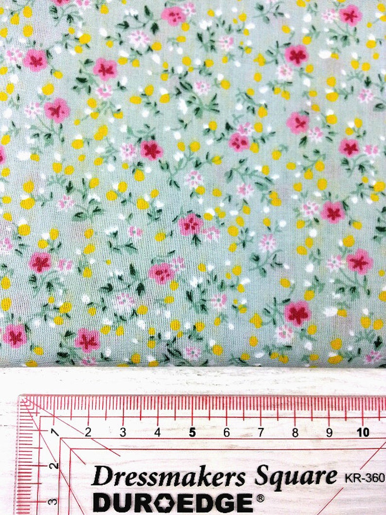 May include: A close-up of a light green fabric with a small floral print. The fabric has tiny yellow and pink flowers with green leaves. A ruler is visible at the bottom of the image, showing the fabric is 10 inches wide.