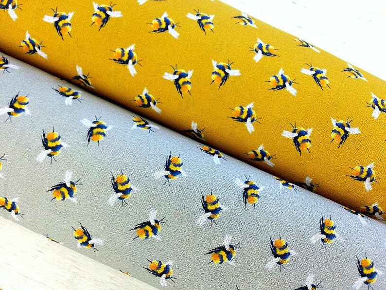 May include: Two rolls of fabric with a repeating pattern of yellow and black bumblebees. The fabric on top is a mustard yellow color and the fabric on the bottom is a light gray color.