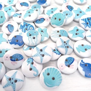 May include: A collection of white wooden buttons featuring a variety of sea creature designs, including starfish, seashells, seahorses, jellyfish, and fish. The buttons are painted in shades of blue and pink.