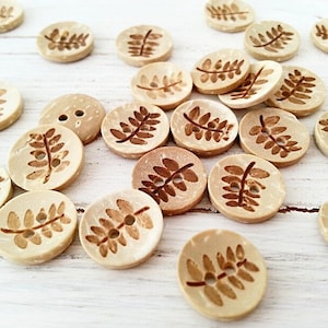 May include: A collection of beige wooden buttons with a brown leaf design. The buttons are scattered on a white surface.