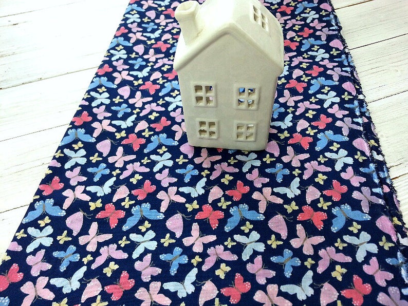 May include: A navy blue fabric with a repeating pattern of pink, blue, and yellow butterflies. A white ceramic house sits on top of the fabric.