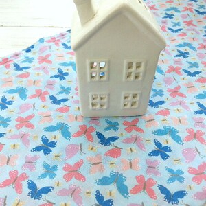May include: A white ceramic house-shaped candle holder sits on a blue fabric with a repeating pattern of pink, blue, and yellow butterflies.