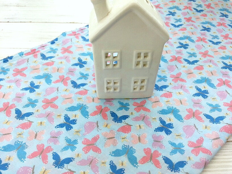 May include: A white ceramic house-shaped candle holder sits on a blue fabric with a repeating pattern of pink, blue, and yellow butterflies.