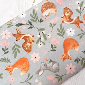 May include: A light blue fabric with a repeating pattern of woodland animals and flowers. The animals include a deer, a fox, a rabbit, a hedgehog, and a squirrel. The flowers are pink and white.