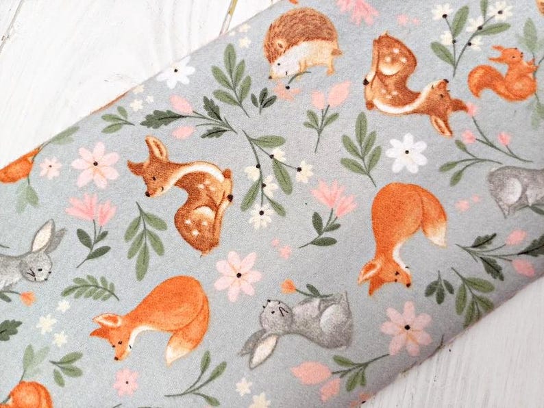 May include: A light blue fabric with a repeating pattern of woodland animals and flowers. The animals include a deer, a fox, a rabbit, a hedgehog, and a squirrel. The flowers are pink and white.
