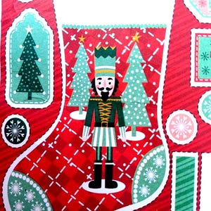 Op de afbeelding: Een kerstsokdecoratie met een centrale notenkrakerfiguur in een groen uniform, staand tussen twee lichtgroene dennenbomen. De sok heeft een rood geruite achtergrond met witte sneeuwvlokaccenten en een witte boord aan de bovenkant.