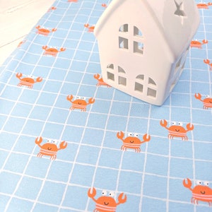 Op de afbeelding: Een lichtblauwe stof met een wit rasterpatroon en oranje krabben. Een wit keramiek huis met uitgesneden ramen staat op de stof.