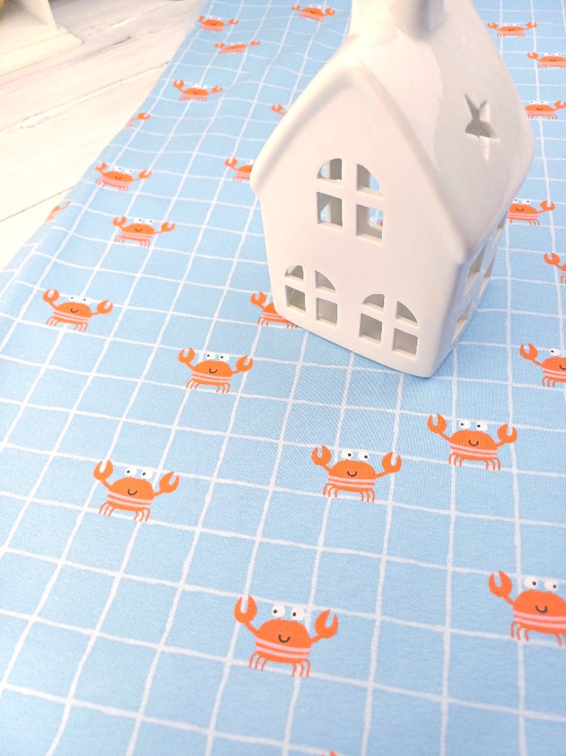 Op de afbeelding: Een lichtblauwe stof met een wit rasterpatroon en oranje krabben. Een wit keramiek huis met uitgesneden ramen staat op de stof.