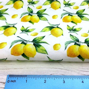 May include: White fabric with a repeating pattern of yellow lemons and green leaves. The fabric is folded over a wooden surface with a ruler measuring 5 inches.