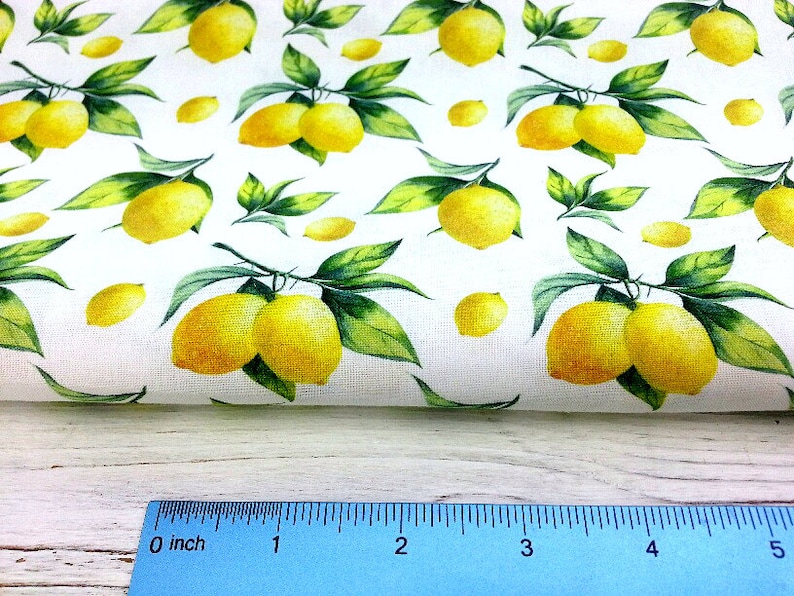 May include: White fabric with a repeating pattern of yellow lemons and green leaves. The fabric is folded over a wooden surface with a ruler measuring 5 inches.