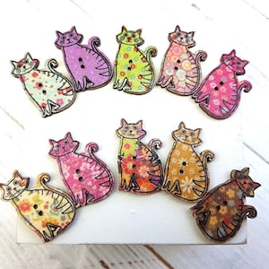 May include: Nine colourful wooden cat buttons with floral patterns. The buttons are approximately 5 cm in diameter and have two holes for sewing.