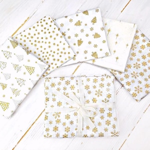 May include: White fabric squares with gold Christmas tree, snowflake, and star patterns. One bundle is tied with a white ribbon. The fabrics are arranged on a white wooden surface.