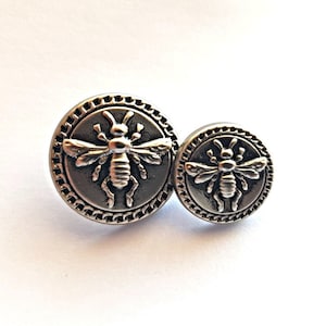 May include: Two silver metal buttons featuring a bee design. The larger button is slightly bigger than the smaller button.