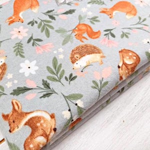 May include: A gray fabric with a repeating pattern of woodland animals and flowers. The animals include deer, foxes, squirrels, and hedgehogs. The flowers are pink, white, and green.