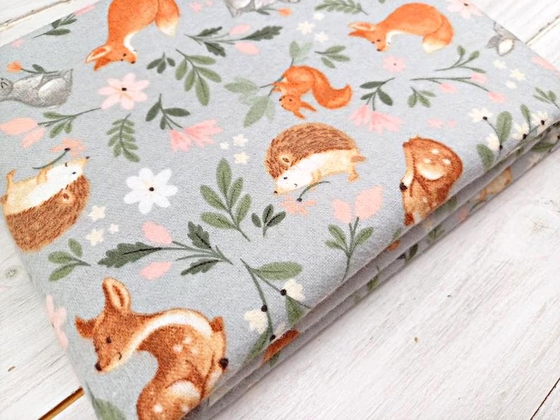 May include: A gray fabric with a repeating pattern of woodland animals and flowers. The animals include deer, foxes, squirrels, and hedgehogs. The flowers are pink, white, and green.