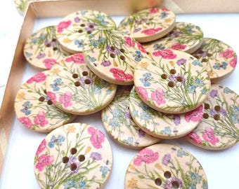 May include: A pile of wooden buttons with a floral print. The buttons are cream coloured with pink, blue, and yellow flowers. The buttons have four holes.