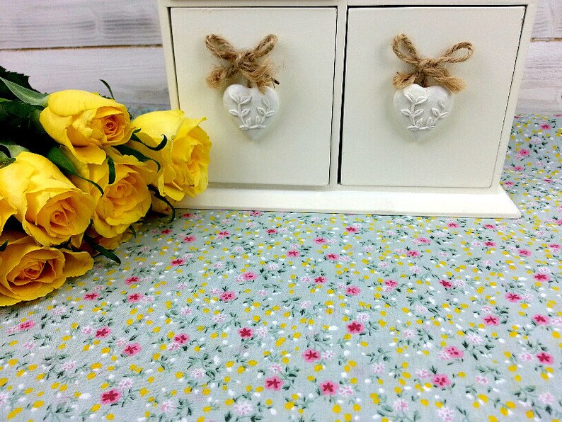 May include: A white wooden box with two drawers, each with a heart-shaped knob and a bow made of twine. The box is sitting on a light blue fabric with a floral pattern. A bouquet of yellow roses is in the foreground.