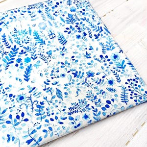 Op de afbeelding: Een gevouwen stuk stof met een witte achtergrond en een herhalend patroon van blauwe bloemen en botanische elementen. Het ontwerp bevat bladeren, bloemen en stelen in verschillende tinten blauw, waardoor een aquareleffect ontstaat. De stof ligt op een wit houten oppervlak.
