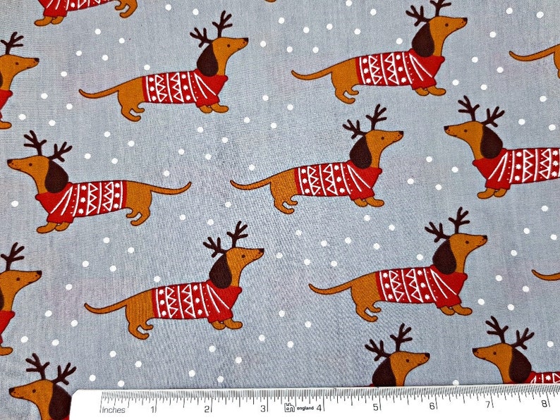 May include: A gray fabric with a white polka dot pattern featuring a repeating design of brown dachshunds wearing red and white reindeer sweaters.