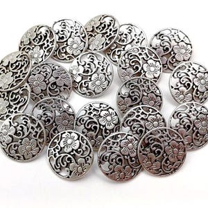 Könnte beinhalten: Eine Sammlung silberfarbener Metallknöpfe mit filigranen Blumendesigns. Die Knöpfe sind rund und haben einen erhöhten Rand.