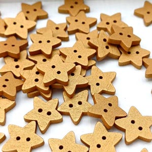 May include: A pile of star-shaped wooden buttons in a golden hue. Each button features two holes for stitching. The buttons are scattered on a white surface, creating a textured and visually appealing arrangement.