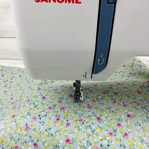 May include: A white sewing machine with the brand name "JANOME" printed on the front. The machine is set up to sew a piece of light green fabric with a small pink and yellow floral pattern.