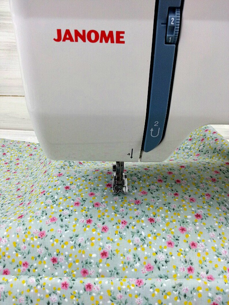 May include: A white sewing machine with the brand name "JANOME" printed on the front. The machine is set up to sew a piece of light green fabric with a small pink and yellow floral pattern.
