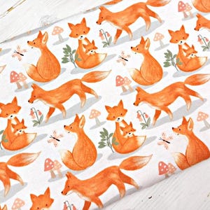 May include: White fabric with a repeating pattern of orange foxes. The foxes are depicted in a cartoon style, with some holding a smaller fox cub. The background is white with small pink and white mushrooms and butterflies.