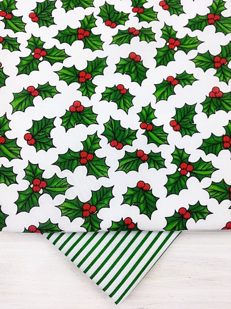 May include: White fabric with a repeating pattern of green holly leaves with red berries. A green and white striped fabric is folded on top of the holly fabric.