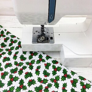 May include: A white sewing machine with the brand name "JANOME" printed in red on the side. The machine is positioned over a white fabric with a repeating pattern of green holly leaves and red berries.