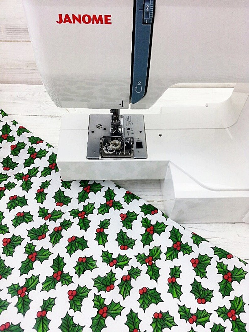 May include: A white sewing machine with the brand name "JANOME" printed in red on the side. The machine is positioned over a white fabric with a repeating pattern of green holly leaves and red berries.