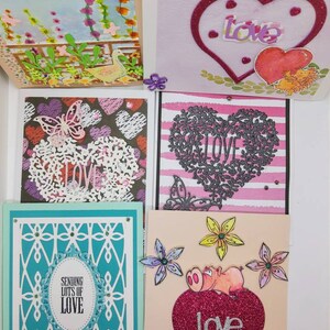 May include: Five handmade greeting cards with different designs featuring hearts, flowers, butterflies, and the word "love". The cards are all decorated with glitter and other embellishments.