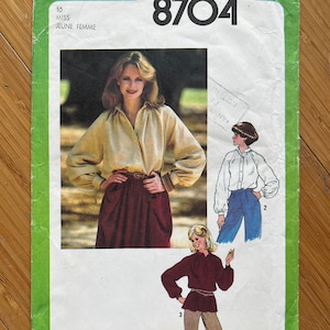 Può includere: Modello di cucito Simplicity numero 8704 per una signorina o una giovane donna. Il modello include tre stili diversi di camicette a maniche lunghe. La modella indossa una camicetta bianca con colletto e una gonna marrone. Il modello è stampato su uno sfondo verde.