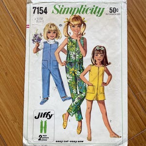May include: Vintage Simplicity sewing pattern number 7154 for girls' clothing. The pattern includes three different styles: a blue jumpsuit, a green floral print jumpsuit, and a yellow jumpsuit. The pattern is labelled "Jiffy" and "easy cut-easy sew".