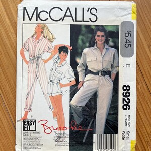 May include: McCall's pattern 8926 for a jumpsuit, labelled "Easy Fit" and "Small Petite." The pattern features a white jumpsuit with pink stripes, a white jumpsuit with a blue belt, and a white jumpsuit with a black belt. The pattern is signed "Brooke Shields."
