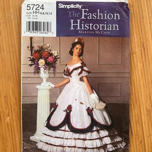 May include: A Simplicity 5724 sewing pattern envelope for The Fashion Historian by Martha McCain. The cover features a person wearing a white ball gown with red and black floral accents, a ruffled skirt, and white gloves, standing next to a pedestal.