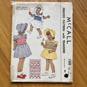 Op de afbeelding: Vintage McCall's naaipatroon voor een kindersmockjurk en -muts. Het patroon is gedrukt op papier en bevat instructies voor het maken van de jurk en de muts. Het patroon is voor maten 2, 4 en 6. Het patroon is geel en blauw.