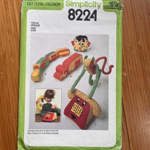 May include: A Simplicity 8224 sewing pattern envelope featuring illustrations of stuffed fabric toys including a telephone, a clock, a train, and a truck. The green and white paper envelope displays the text Pattern/Patron, Simplicity 8224, and One Size.