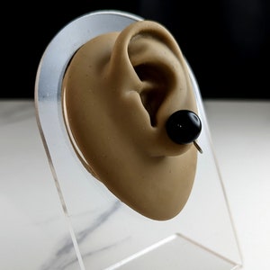 May include: A black ear stud with a silver post, displayed on a clear plastic ear model.