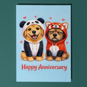 May include: A greeting card with a light blue background features two dogs dressed in panda and red panda costumes. The panda dog is smiling, while the red panda dog has a serious expression. The words "Happy Anniversary" are written in red.