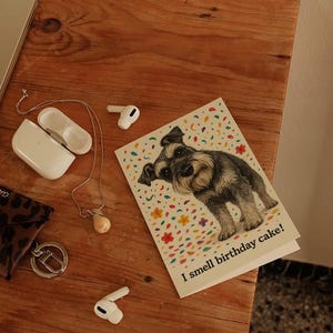 May include: A greeting card featuring a Schnauzer dog illustration with the text "I smell birthday cake!" on a white background with colorful confetti. The card is on a wooden surface with other items.