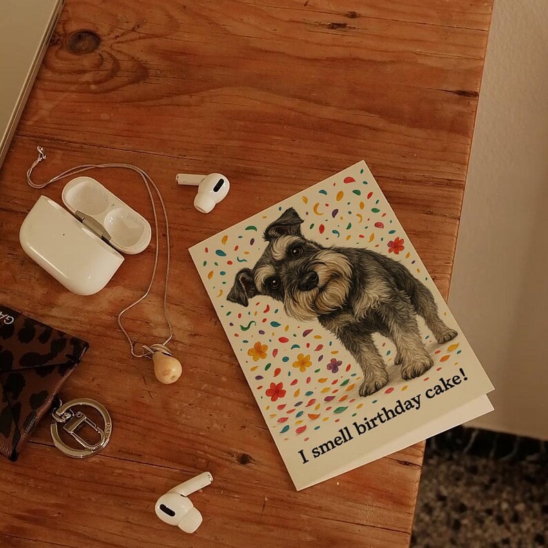 May include: A greeting card featuring a Schnauzer dog illustration with the text "I smell birthday cake!" on a white background with colorful confetti. The card is on a wooden surface with other items.
