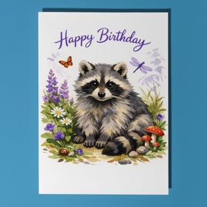 Op de afbeelding: Verjaardagskaart met een aquarel illustratie van een wasbeer omringd door bloemen, paddenstoelen en een slak. De kaart heeft de tekst "Happy Birthday" in paars schrift. Een vlinder en een libelle zijn ook in het ontwerp opgenomen.