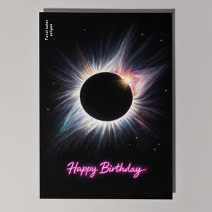 May include: A birthday card featuring a total solar eclipse illustration. The design shows a black disc representing the moon, encircled by a vibrant corona against a black backdrop. The words "Happy Birthday" are written in pink neon.