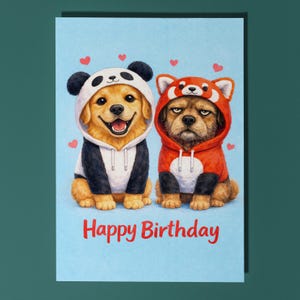May include: A birthday card featuring two dogs dressed in animal costumes. One dog is in a panda outfit, the other in a red panda costume. The words "Happy Birthday" are written in red on a light blue background, with small pink hearts.