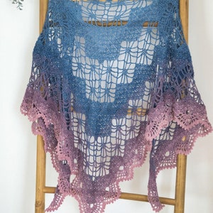 Pode incluir: Um xale de crochê com um degradê de cores rosa e azul. O xale tem um delicado padrão de renda e uma borda festonada.